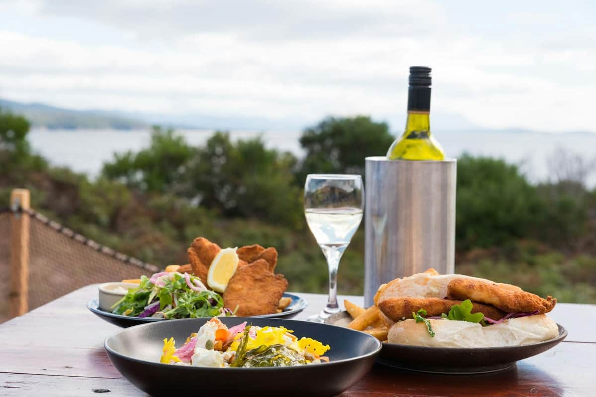 Bruny Island: Guided Food Tour from Hobart + Ferry Ride + Bligh Museum Entry A table set with plates of food, bread, fried items, salad, a wine bottle in a cooler, and a glass of white wine.