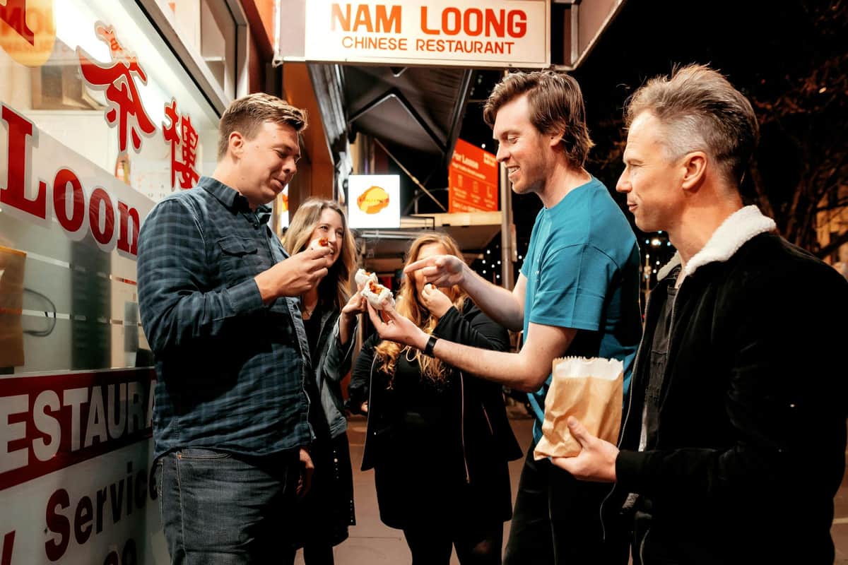 A group of five people standing outside a Chinese restaurant at night, sharing food and talking under bright signage.