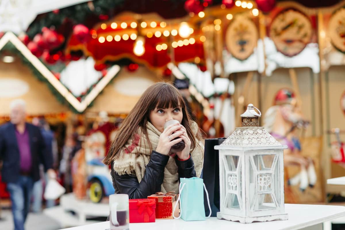 Woman sipping a drink at an outdoor holiday market, surrounded by gift bags and a lantern, with festive decorations in the background.