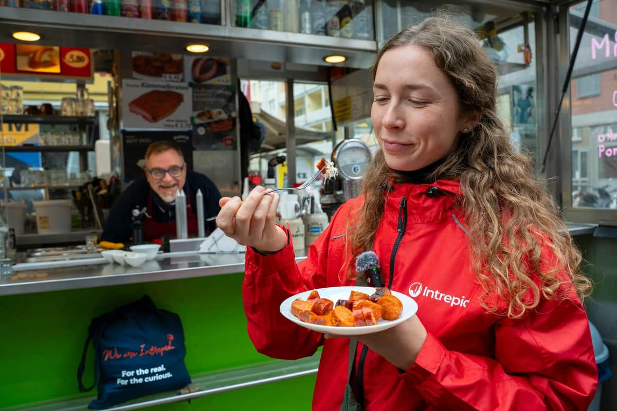 A woman in a red jacket enjoys a plate of food in front of a food stand, with a man smiling in the background.