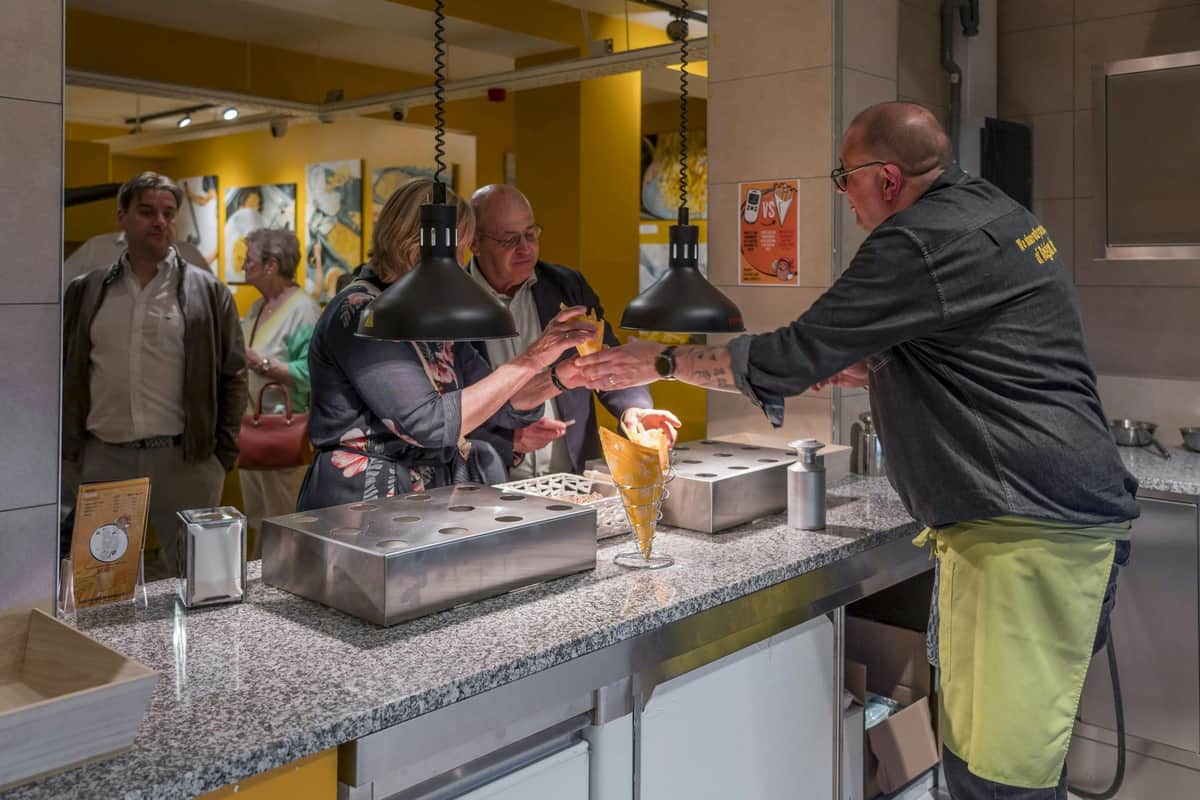 People waiting at a counter; a man serves cones with food to a woman under hanging lights in a brightly lit room.