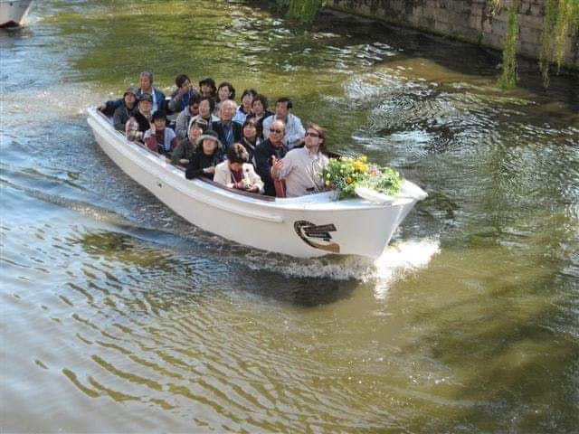 A group of people seated in a white boat cruising along a canal. A floral arrangement is at the front of the boat.
