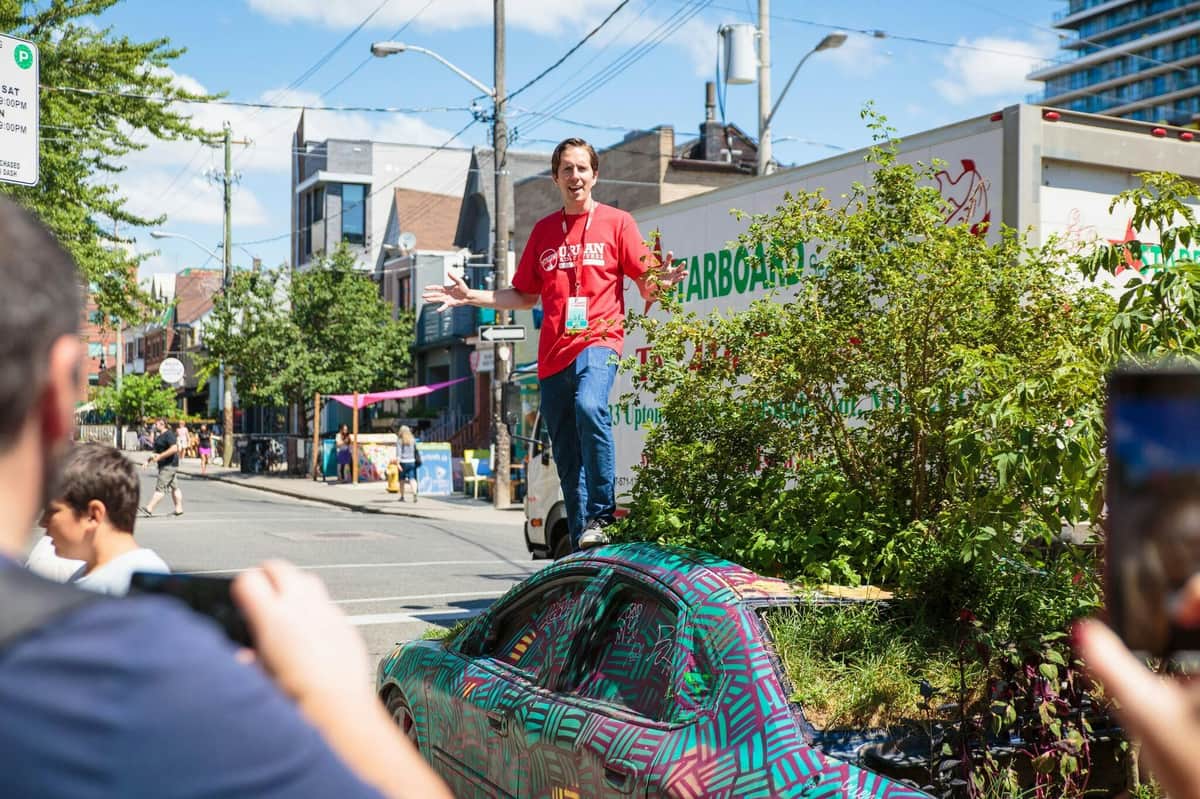 A man in a red shirt and jeans stands on a creatively painted car surrounded by greenery on a city street.