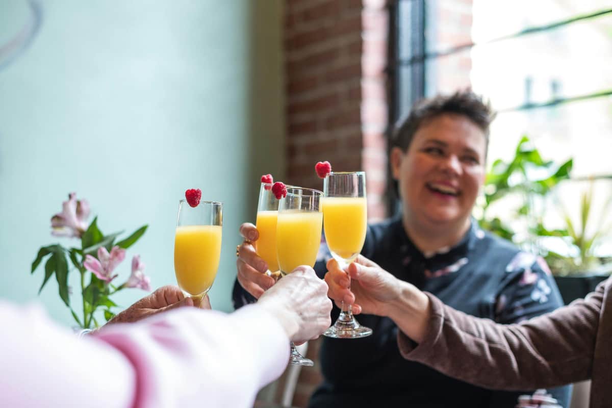 Vancouver: Gastown Guided Brunch Tour Four people clink glasses of orange drinks with raspberries. A person in the background is smiling, and there are plants near a window.