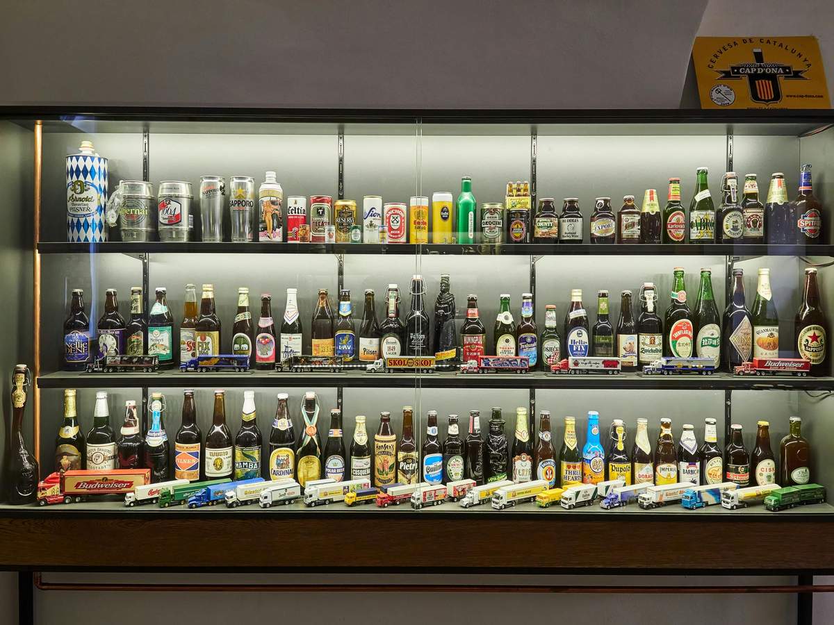 Prague Beer Tour & Czech Beer Museum A display case with three shelves containing various bottles and cans of beer, along with miniature trucks at the bottom.