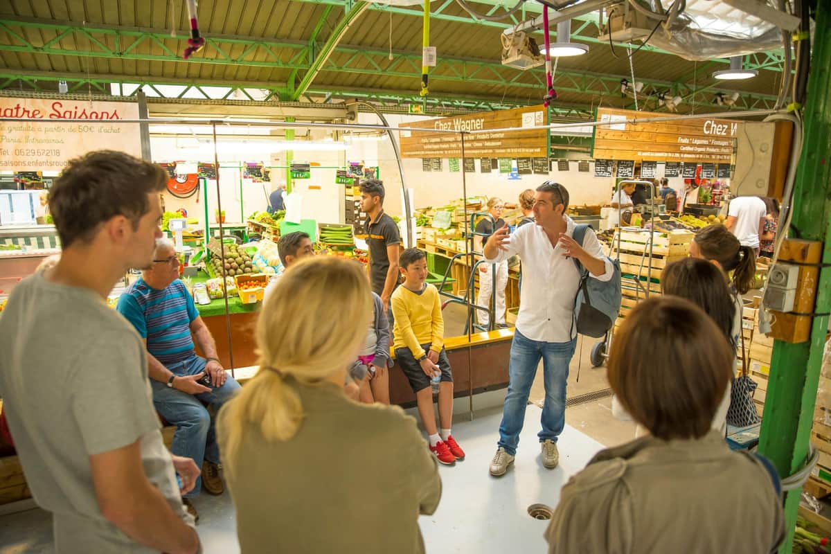 A group of people gathered around a man speaking in a market with fresh produce and stalls visible in the background.