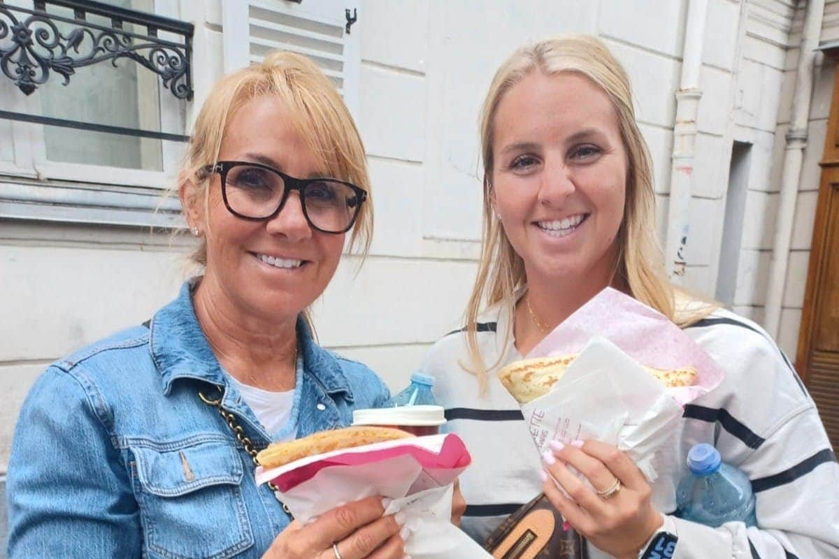 Dos mujeres, sonrientes y con comida envuelta en papel en las manos, de pie fuera, cerca de un edificio blanco.