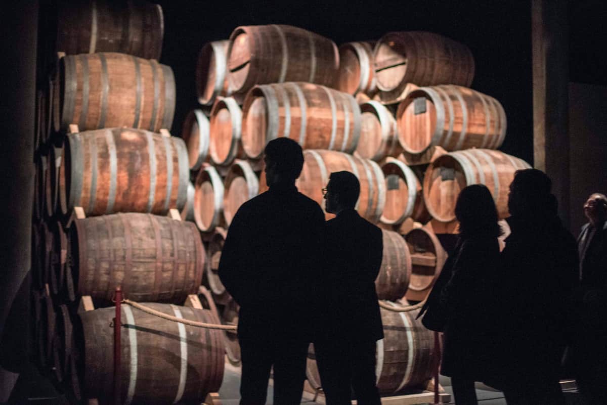 Calvados Experience Three individuals observe stacks of large wooden barrels in a dimly lit setting.