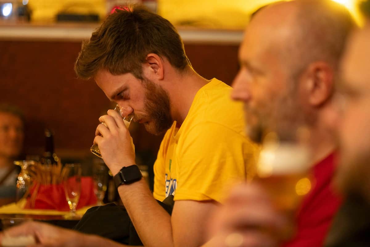 A man in a yellow shirt sniffs a drink glass while sitting indoors. Another person is partially visible in the foreground.