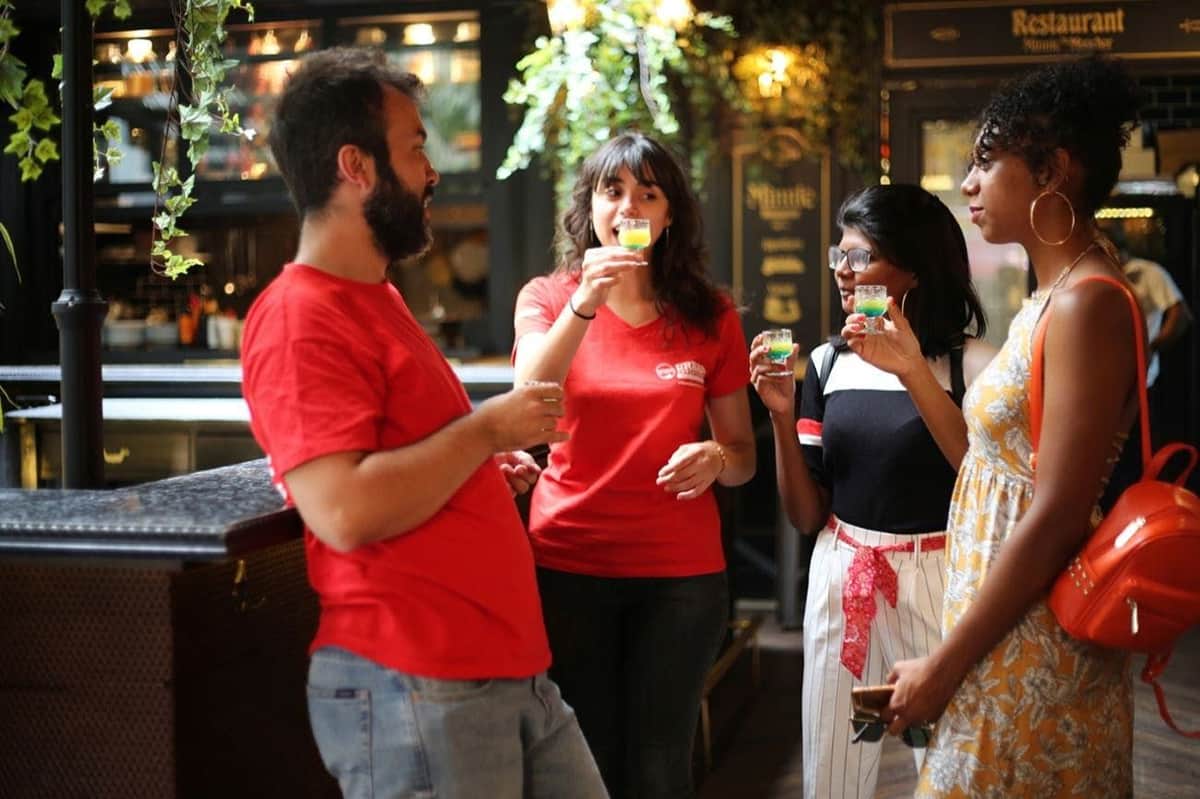 Athens: Small Group Guided Night Tour + Snacks & Drinks Four people stand together in a social setting, holding small glasses and engaging in conversation.