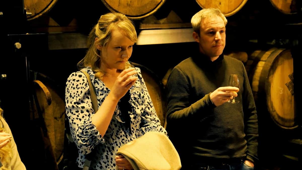Dublin's Distillery Trail: VIP Private Guided Tour Two people are sampling drinks in front of wooden barrels. The woman holds a glass and a coat, while the man holds his glass and looks ahead.