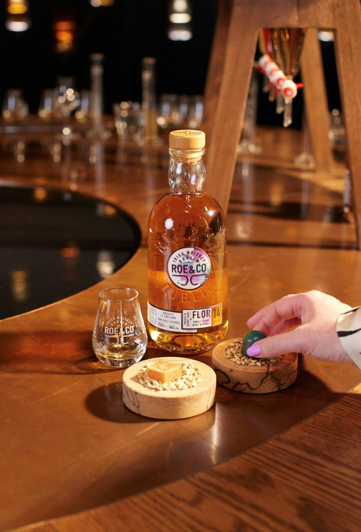 Roe & Co Distillery: Beyond the Blend + Signature Cocktail A bottle of Roe & Co whiskey beside a branded glass and two wooden dishes, one with a sugar cube and another with seeds, placed on a wooden table.