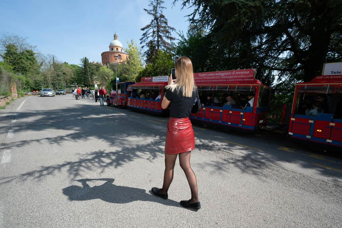 A woman in a red skirt photographs a red sightseeing train on a sunny street with trees and a dome-shaped building in the background.