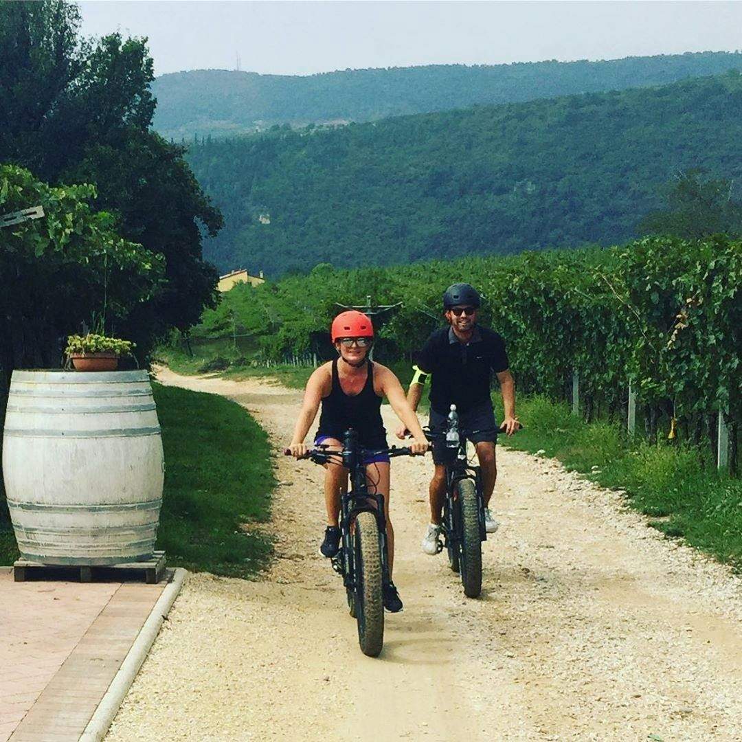 Two people cycling on a gravel path through a vineyard, wearing helmets. Hills and greenery are visible in the background.