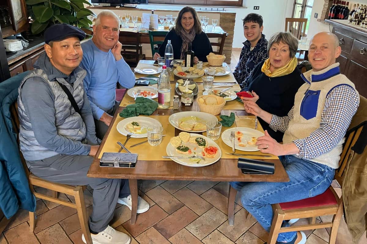 Tuscany: Guided Tour + Lunch & Drinks A group of six people seated at a table with plates of food, glasses, and bottles, smiling in a casual dining setting.