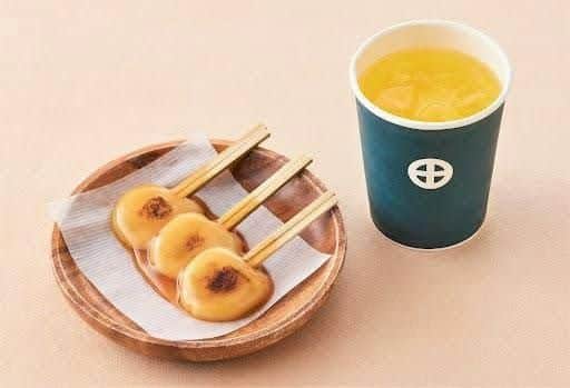 Three skewers of glazed grilled dumplings on a wooden plate next to a cup of yellow beverage with ice.