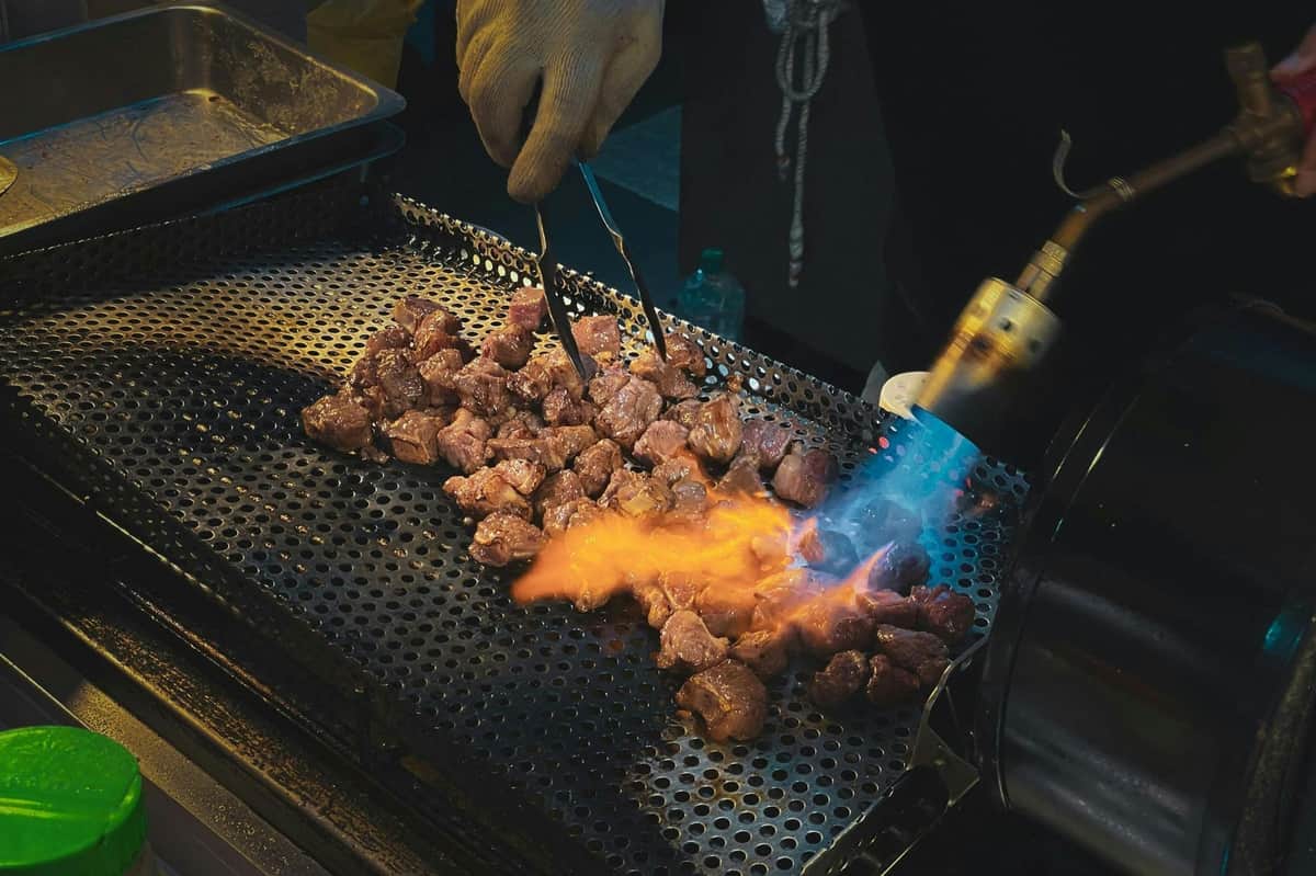 A person sears chunks of meat on a perforated grill using tongs and a blowtorch with a visible flame.