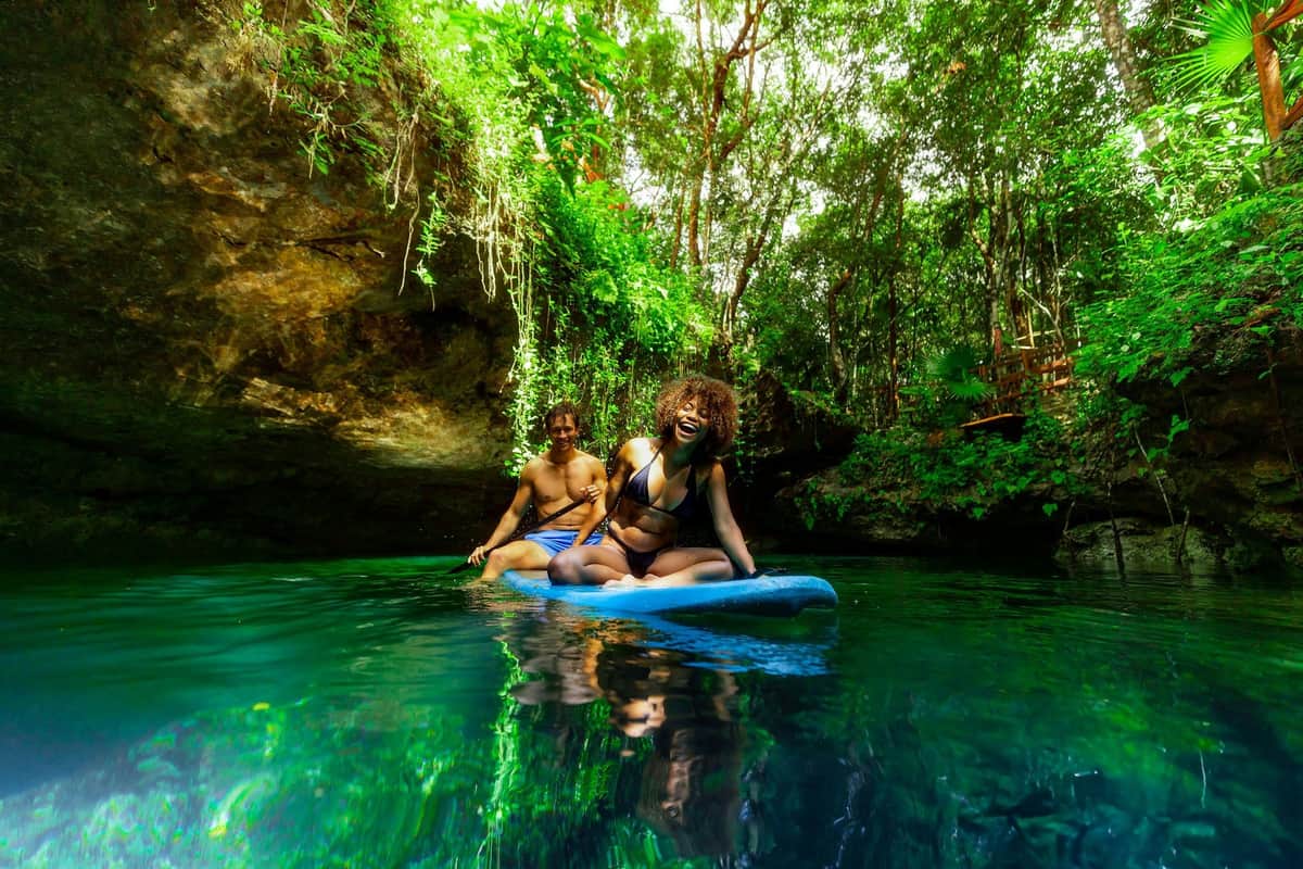 Cancun: Cenotes Adventure + Tequila Tasting + Lunch Two people enjoy paddleboarding on clear water surrounded by lush greenery and rocky cliffs in a natural setting.