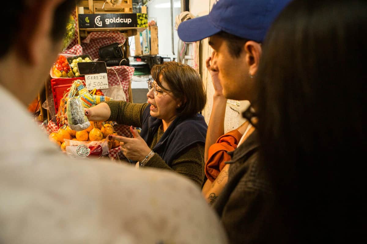 Heart of Mexico City: Flavors of Our Heritage A woman in a market stall interacts with customers, displaying fruits and wearing glasses, while others observe or select items.