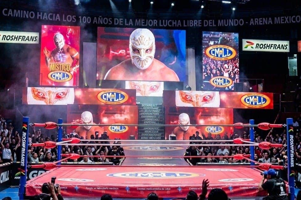 A wrestling ring surrounded by an audience, with large screens displaying a masked wrestler and CMLL logos.