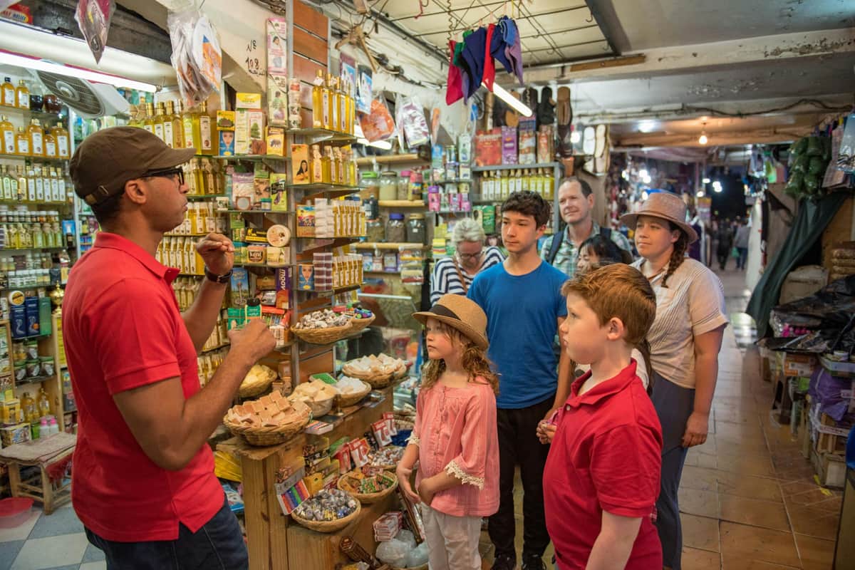 Marrakesh: Guided Food Tour + Dinner A group of people, including children, stand attentively in a colorful, crowded market shop as a vendor talks to them.