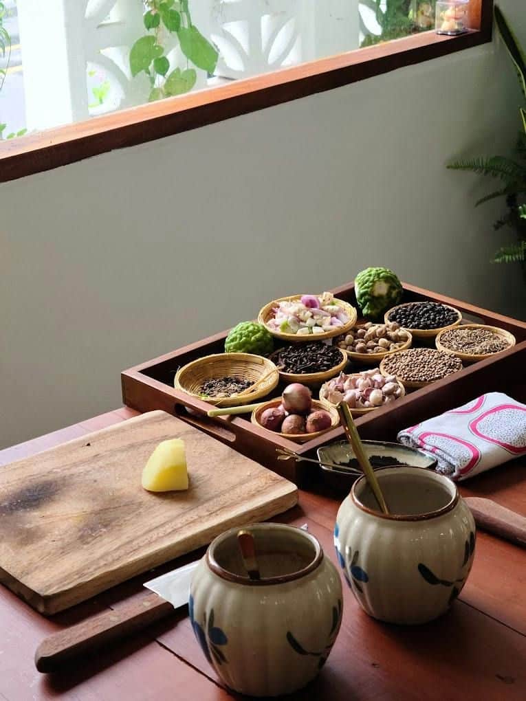 Bangkok : cours de cuisine l'après-midi avec visite du marché et balade en tuk-tuk (Bangkok) Un plateau en bois sur lequel sont disposés divers épices et herbes aromatiques, une planche à découper sur laquelle repose un aliment, ainsi que des bocaux en céramique posés sur un plan de travail en bois.