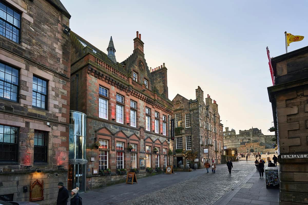 Historic stone buildings line a cobblestone street with a few pedestrians walking and a castle visible in the distance.