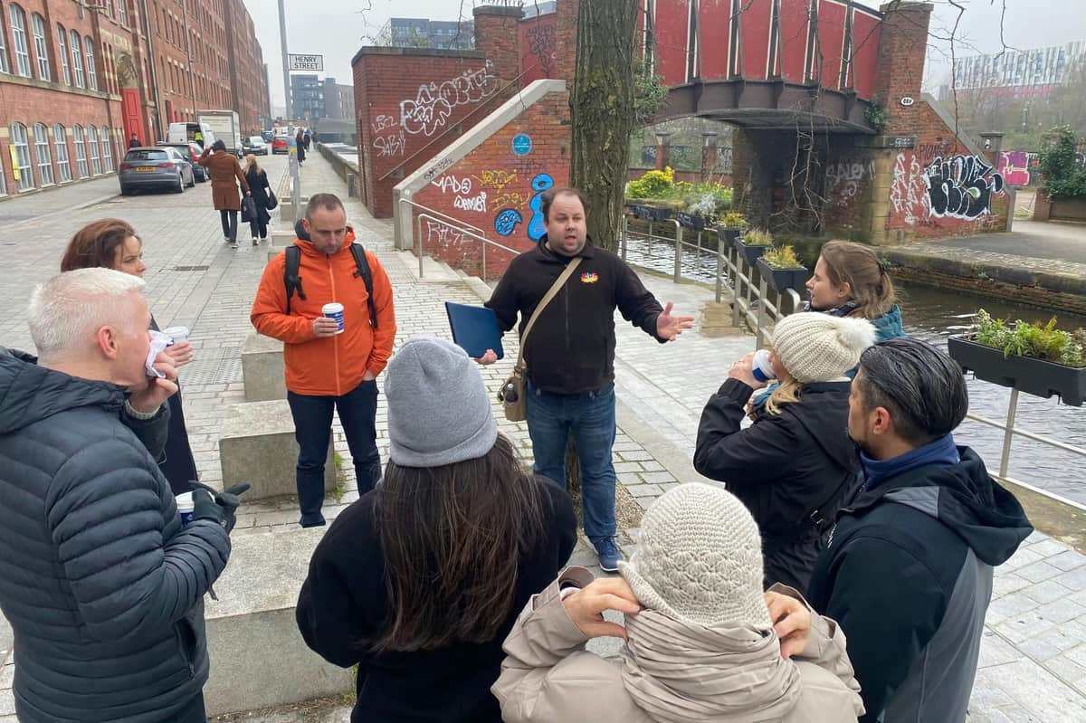 Un homme s'adresse à un petit groupe de personnes dans une rue couverte de graffitis, près d'un canal, avec un pont rouge en arrière-plan.