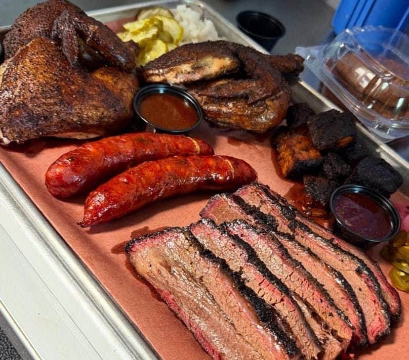 A platter of barbecue meats including brisket, sausage, chicken, and burnt ends, served with pickles and sauce cups.