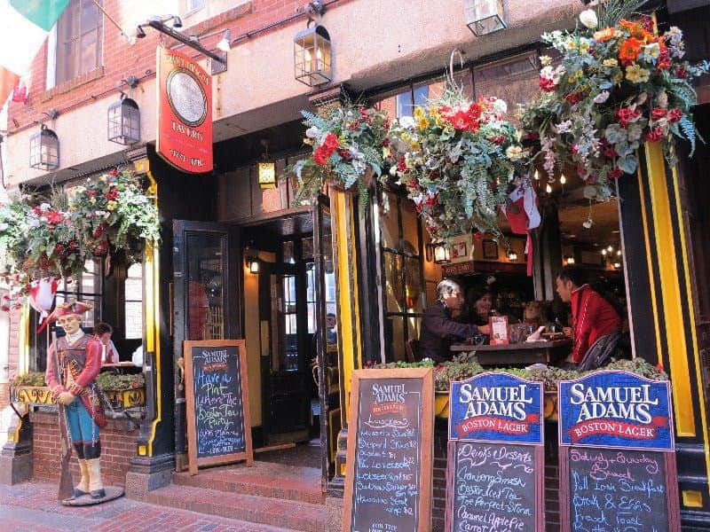 Historisches Boston: Tavernen-Tour Ein lebhafter Pub-Eingang mit Blumendekor, Speisekarten auf Kreidetafeln und sitzenden Gästen im Inneren. Die Werbung für Samuel Adams ist sehr auffällig.