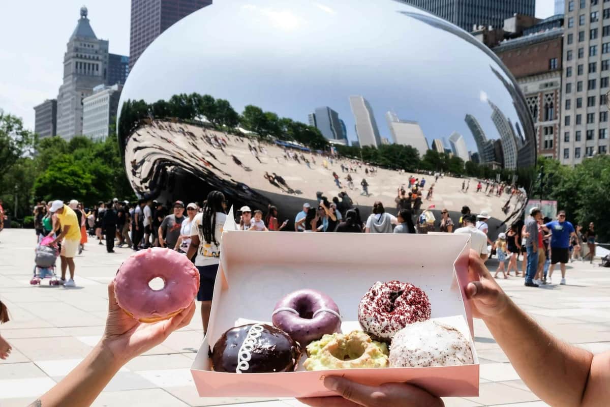 Chicago: Downtown Guided Donut Tasting Tour Person mit einem rosa Donut und einer Schachtel mit verschiedenen Donuts vor der reflektierenden Cloud Gate-Skulptur auf einem belebten Platz.