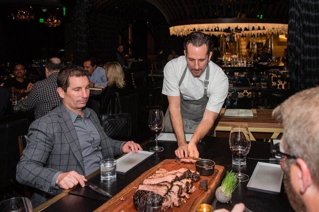 Las Vegas Strip: Guided Steakhouse Food Tour + Transfers A man seated at a restaurant table with sliced meat on a wooden platter, a chef explaining the dish to him.