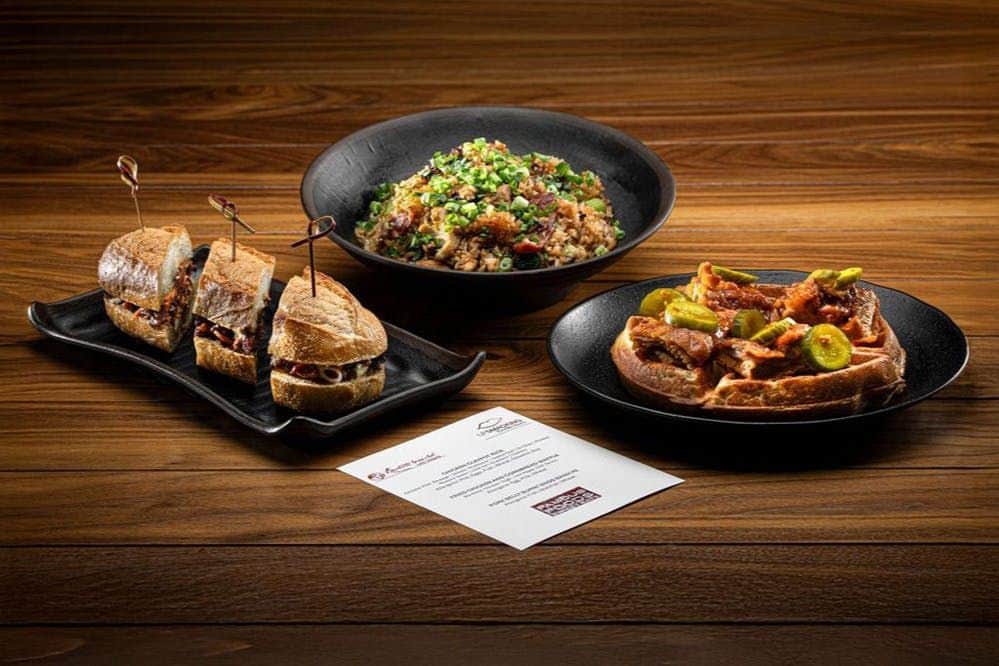 Resorts World Las Vegas: Guided Cocktail Crawl + Food Tastings A wooden table with three dishes of food: a bowl of rice with toppings, sandwiches, and a plate of sausages with pickles.