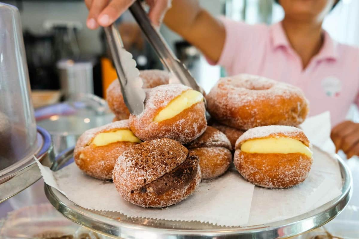 South Beach Miami: Guided Donut Tasting Tour A hand with tongs picks a cream-filled donut from a pile of various filled donuts dusted with sugar on a tray.