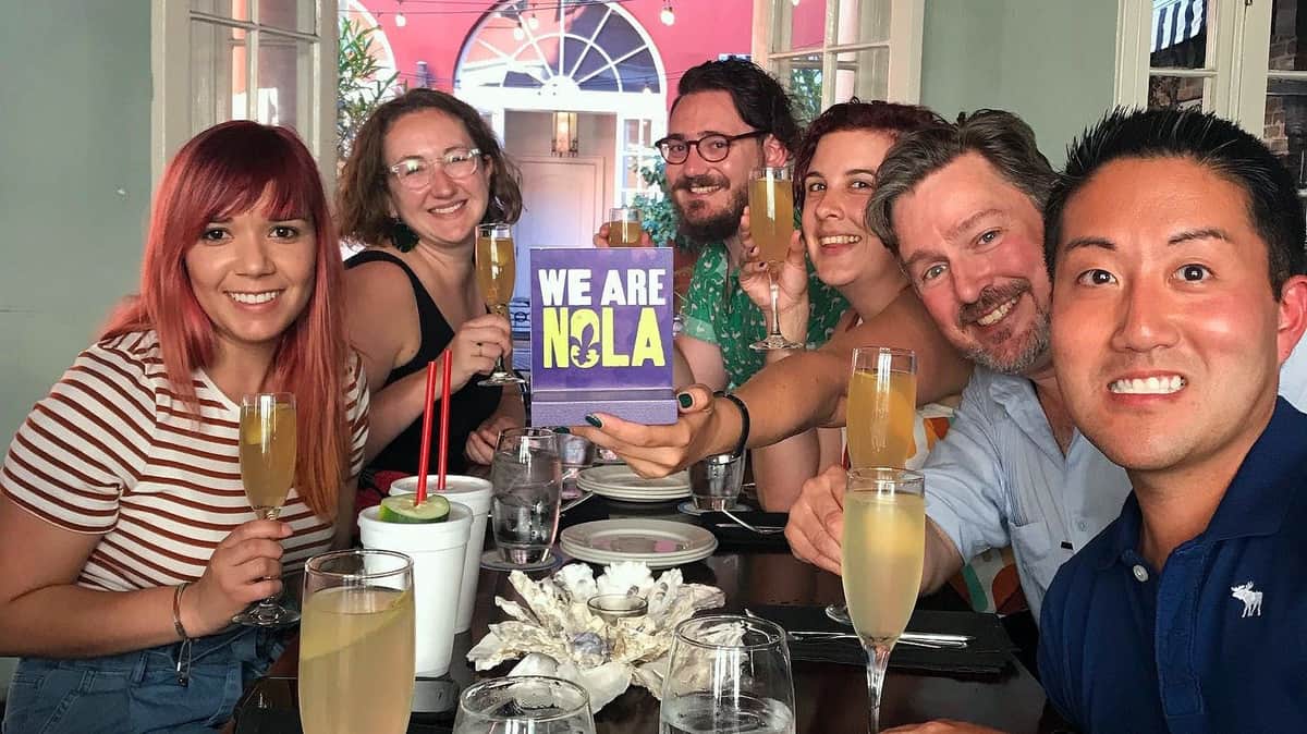 New Orleans: Cocktail and Food History Tour A group of people at a table holding drinks, with one holding a sign reading "We Are NOLA," in a festive setting.