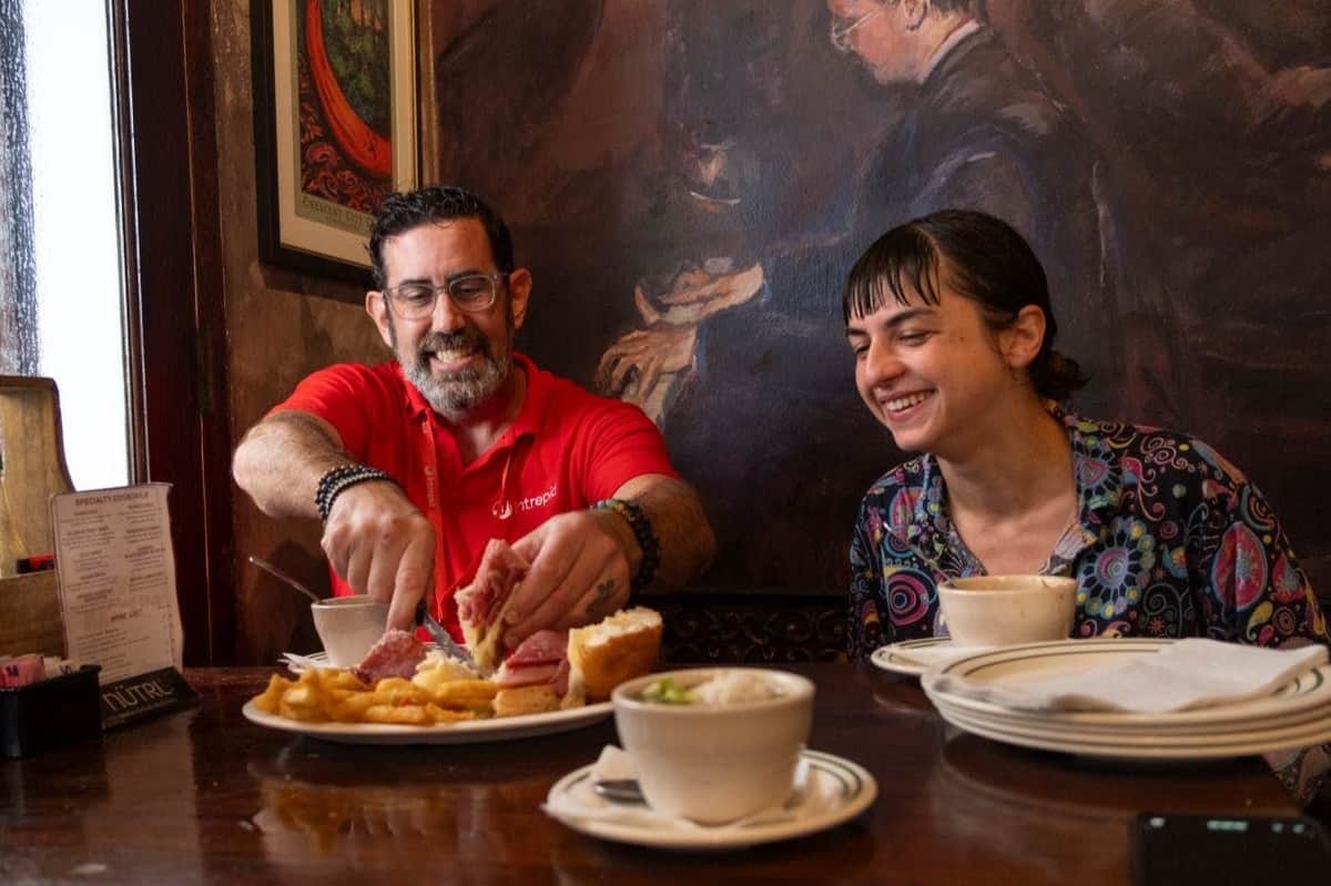 New Orleans: French Quarter Guided Food Tour + 5 Tastings A man and woman enjoy a meal at a rustic restaurant, sharing sandwiches and fries, with coffee cups and a painting in the background.