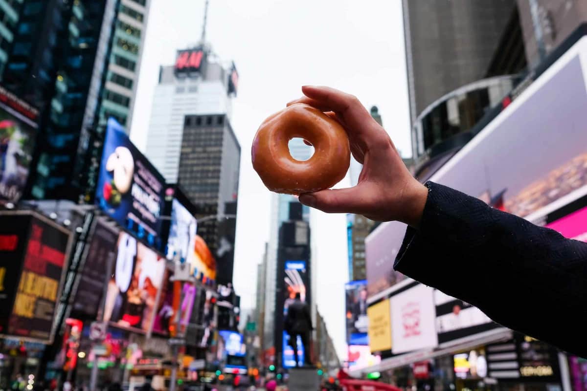 New York: Times Square Guided Tour + Donut Tasting A hand holding a glazed donut in the foreground with Times Square's bright billboards and tall buildings in the background.