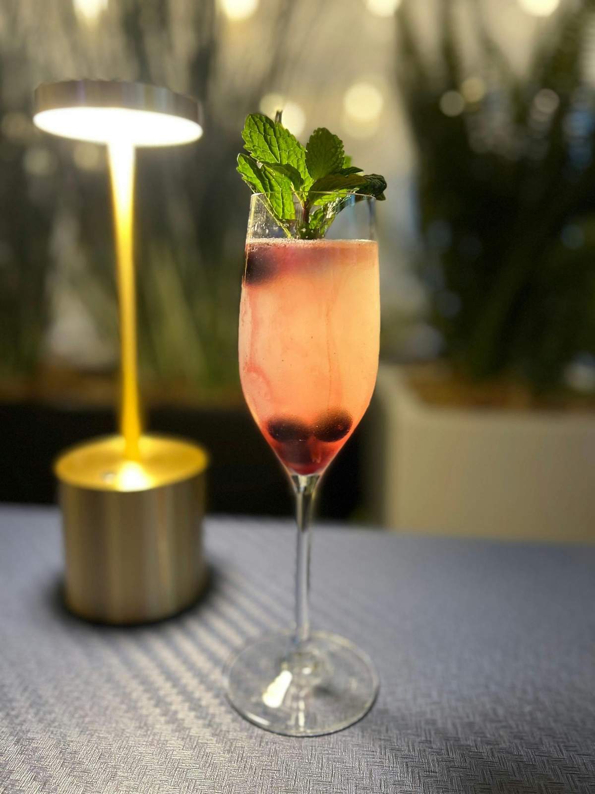 Craft Cocktail Gourmet Food Tour A pink cocktail in a flute glass garnished with mint leaves and berries, placed on a table near a warm-lit lamp.
