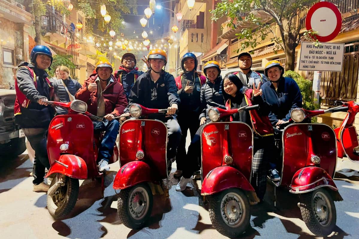 Group of people in helmets posing with red scooters on a brightly lit street at night.
