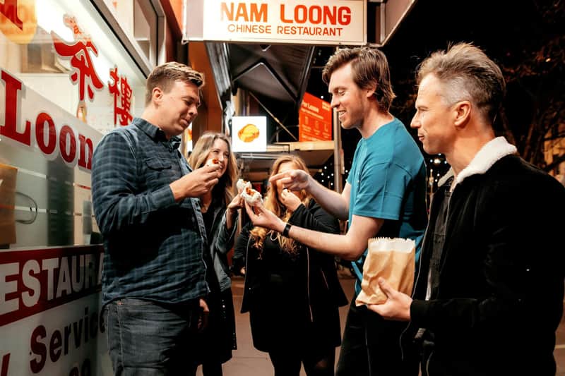 Melbourne: Guided Dinner Tour + Drink A group of five people standing outside a Chinese restaurant at night, sharing food and talking under bright signage.