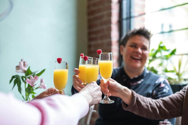 Four people clink glasses of orange drinks with raspberries. A person in the background is smiling, and there are plants near a window.
