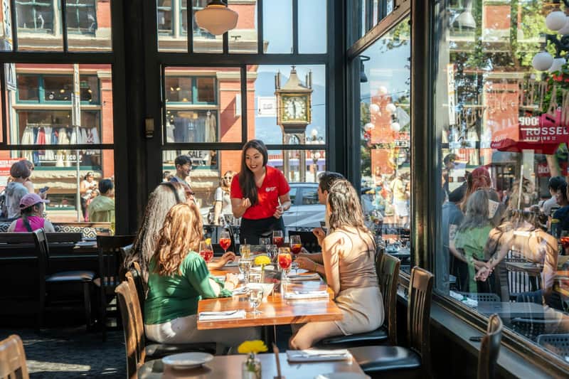 A group of people seated at a restaurant table being served by a waitress. Large windows and a street view are in the background.