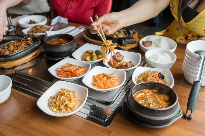 A spread of Korean Barbeque side dishes on a table with a guest picking up vegetables with a chopstick