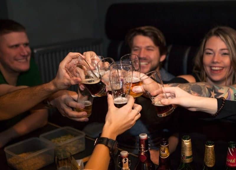 Group of people smiling and clinking beer glasses together, with bottles on a table in the foreground.