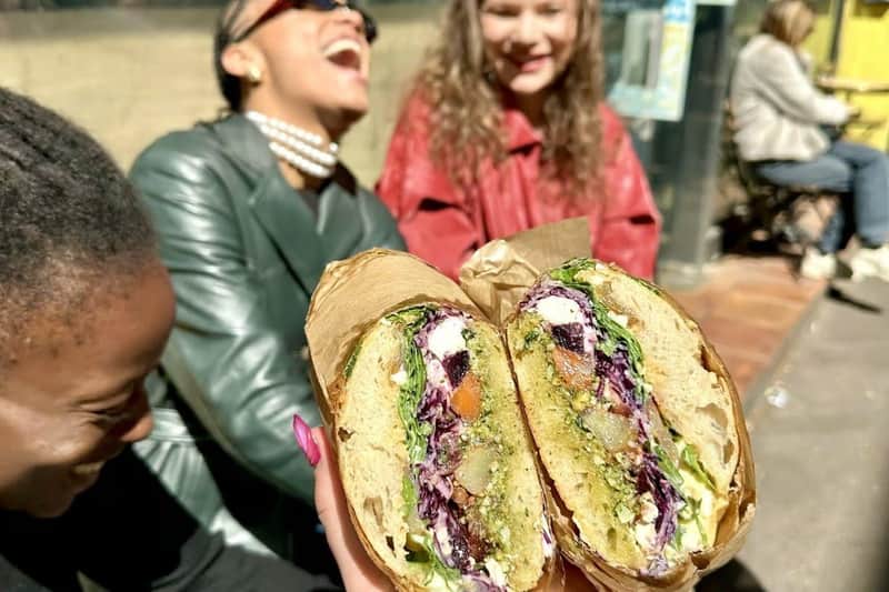 Two sandwiches held in the foreground and 3 women laughing in the background in front of a restaurant