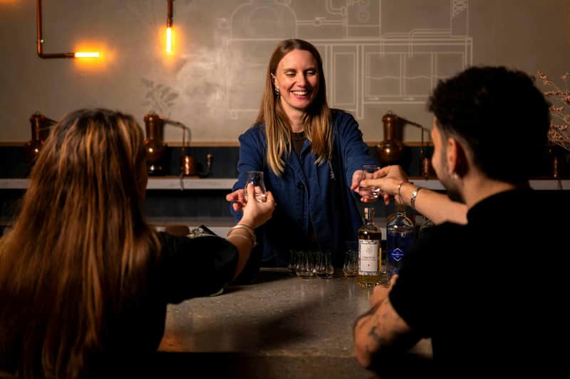 Una mujer sonriente detrás de un mostrador entrega vasos a dos personas en un entorno cálidamente iluminado con botellas y equipos de destilería.