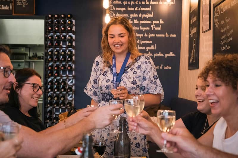 Una mujer sonríe mientras sostiene una copa de vino, rodeada de otras personas que brindan con sus copas en un ambiente acogedor con botelleros.