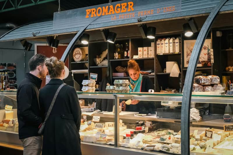 Hay una persona de pie en el mostrador de una tienda de quesos con el letrero «FROMAGER», mientras el dependiente prepara el queso detrás de la vitrina de cristal.