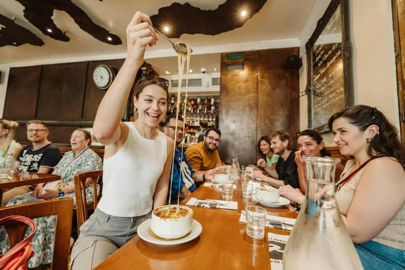 Una persona sacando queso fundido de un plato, junto a un grupo de gente sentada a la mesa en un restaurante con un ambiente acogedor.