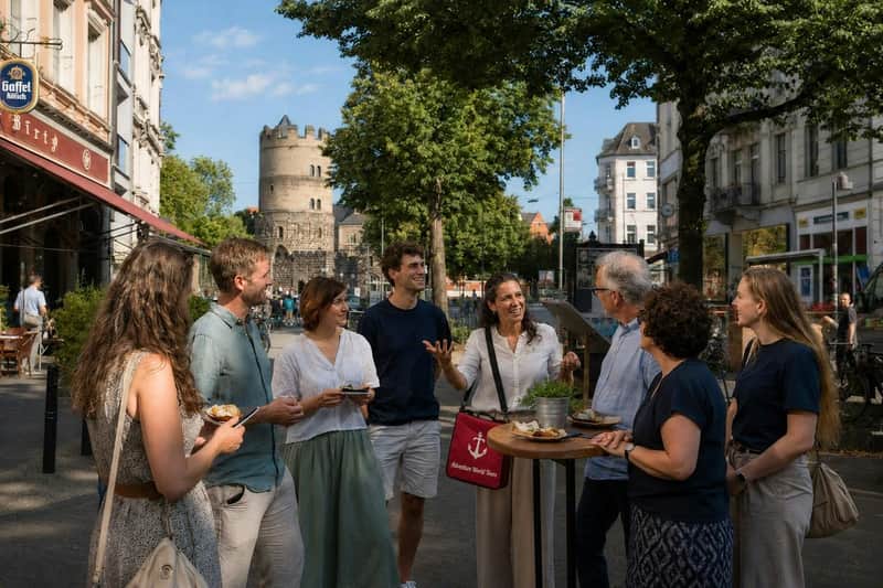 A group of people standing outdoors around a small table, talking and eating, with historic buildings and trees in the background.